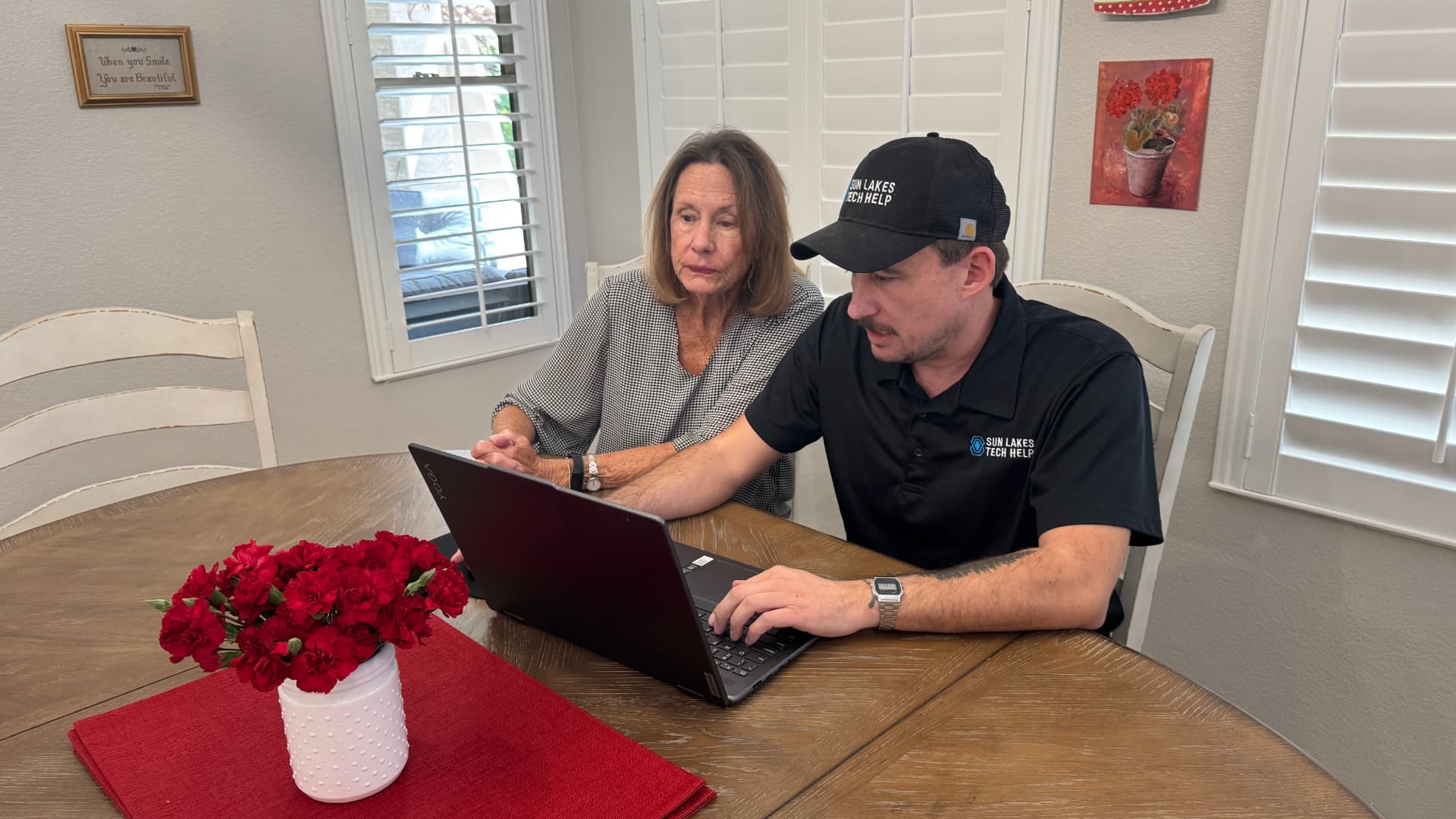 A Sun Lakes Tech Help technician sitting beside a neighbor at her kitchen table, showing her laptop screen